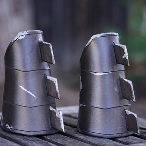 May include: A pair of silver-colored metal gauntlets with a textured surface and sharp edges. The gauntlets are made of multiple pieces of metal that are joined together.