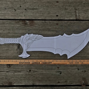 May include: A white paper cutout of a sword with a detailed handle and blade. The sword is approximately 20 inches long and is lying on a wooden surface. The text "SOUTHERN LUMBER COMPANY SAN JOSE, CA 294" is visible on a ruler next to the sword.