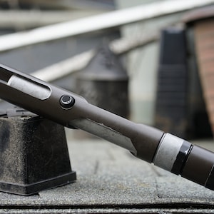 May include: A dark gray lightsaber hilt with a silver accent and a black button. The hilt is resting on a black plastic base.