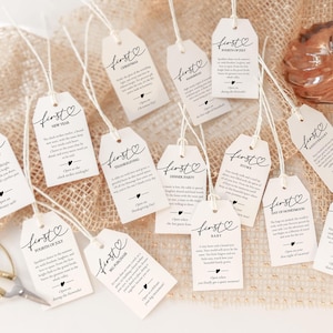 May include: A collection of cream-colored gift tags with the word "first" in elegant script, each with a different occasion like "Valentine's Day" or "Baby." The tags are tied with twine and arranged on a textured surface.