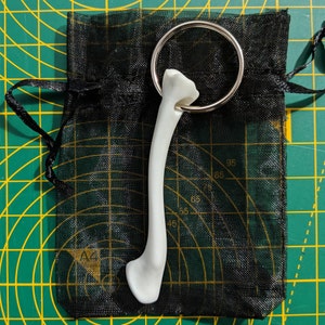 Könnte beinhalten: Ein weißer knochenförmiger Schlüsselanhänger mit einem silbernen Ring. Der Schlüsselanhänger liegt auf einer schwarzen Netz-Tasche.