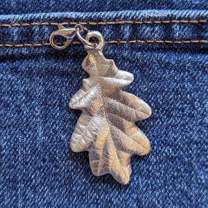May include: A silver oak leaf charm with a silver metal clasp attached to the top. The charm is hanging from a blue denim pocket.