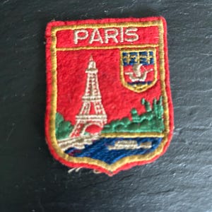 May include: Embroidered patch with the word "PARIS" in gold. The red patch shows the Eiffel Tower, a river, and a coat of arms. The patch has a gold border and is likely a souvenir or travel item, reminiscent of a trip to Paris.