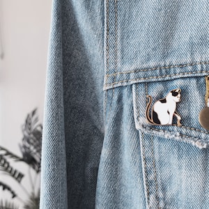 May include: A black and white enamel pin of a cat sitting on a blue denim jacket. The pin is attached to the pocket of the jacket.