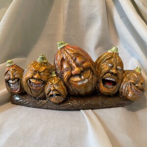 May include: A row of five carved pumpkin faces with toothy grins, arranged on a brown wooden base. The pumpkins are painted in shades of orange, brown, and green.