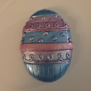 Könnte beinhalten: Ein dekoratives Osterei in Blau-, Rosa- und Silbertönen. Das Ei hat horizontale Bänder mit gepunkteten und gewirbelten Mustern. Das Ei hat eine ovale Form und eine glänzende Oberfläche. Das Ei ist eine festliche Dekoration.