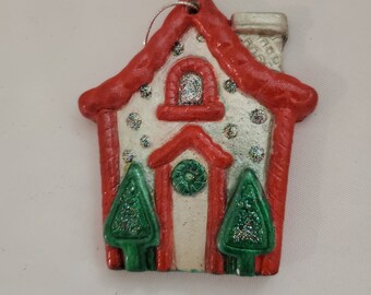 Peut inclure: Ornement de Noël en forme de maison. Le toit et les bordures sont rouges, la façade blanche est ornée de paillettes argentées. Deux sapins de Noël verts et brillants encadrent la porte d'entrée. Une ficelle argentée est attachée pour suspendre.