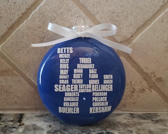 May include: A blue round Christmas ornament with a white ribbon. The ornament has white text listing the names of baseball players, including Seager, Bellinger, Kershaw, and Buehler.