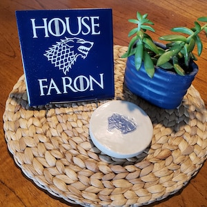 May include: A blue ceramic tile with the words "House Faron" and a white wolf graphic. The tile is on a woven placemat with a white ceramic coaster with a blue fish graphic.