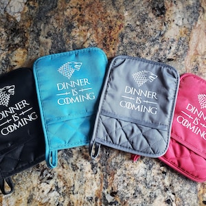 May include: Four oven mitts with a quilted design. Each mitt features a white direwolf graphic and the text "Dinner is Coming". The mitts are in black, teal, grey, and red.