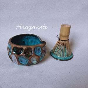 May include: A ceramic matcha bowl with a textured, honeycomb-like exterior featuring shades of turquoise, blue, and brown glaze, paired with a bamboo matcha whisk. The word Aragonite is written in white script above the bowl.