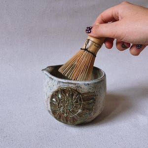 May include: A ceramic matcha bowl with a speckled off-white glaze and a prominent brown ammonite fossil relief on the front. A bamboo matcha whisk is positioned inside the bowl, featuring thin, light-colored bristles tied with black thread.