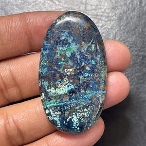 Pode incluir: Uma pedra preciosa polida de formato oval com um padrão complexo de inclusões minerais em azul profundo, turquesa vibrante e castanho terroso. A superfície é lisa e reflexiva, exibindo uma textura natural marmoreada com variada intensidade de cor.
