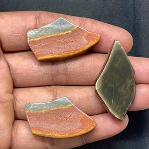 May include: Three polished gemstone slices in shades of red, orange, gray, and brown. The stones have a layered appearance, with a smooth, glossy finish. One stone is a darker green color. The stones are held in a hand.