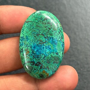 Pode incluir: Gema ovalada com uma vibrante mistura de tons verdes e azuis. A superfície polida revela padrões e texturas intrincados. A gema é segurada em uma mão. Mede aproximadamente 3 cm de comprimento.