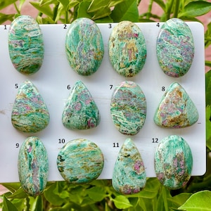 May include: Twelve polished cabochon gemstones arranged in three rows on a white display board. Each stone features a marbled pattern of vibrant green, teal, and white with distinct inclusions of magenta and pink, numbered one through twelve.