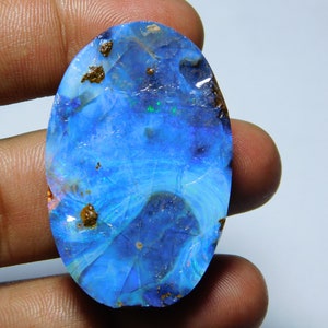 Puede incluir: Un cabujón de ópalo azul con un acabado rugoso y natural. El ópalo tiene un juego de colores azul, verde y blanco. El ópalo está engastado en un anillo de plata.