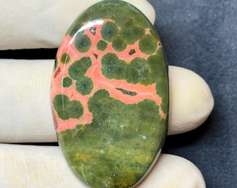 May include: Oval-shaped gemstone with a polished surface. The stone features a green base color with a network of coral-colored veins and dark green spots. The gemstone is held by a gloved hand.