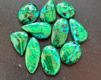 May include: A collection of polished malachite gemstones in various shapes, including oval, teardrop, and irregular forms. The stones display vibrant green hues with swirling patterns and occasional blue-green accents. The gemstones are arranged on a dark surface.