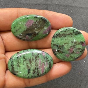 May include: Three polished zoisite gemstones. The stones are oval and round, with a green base color and dark brown and pink inclusions. The gemstones are held in a hand.