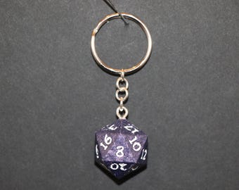 May include: A keychain featuring a dark purple, 20-sided die with white numbering. The die is suspended from a silver-colored chain and keyring. The background is a solid dark gray.