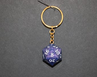 May include: A gold-colored keychain with a blue, twenty-sided die pendant. The die has white numbers and a glittery, speckled finish. The keychain is suspended against a dark background.