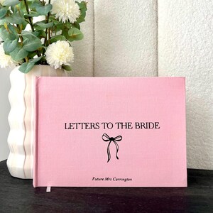 May include: A pink book titled "LETTERS TO THE BRIDE" with a bow illustration. The book has a ribbon bookmark and the text "Future Mrs Carrington" at the bottom. A white vase with flowers is to the left.