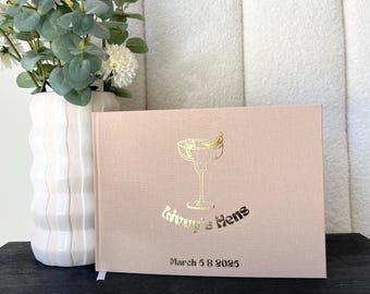 Könnte beinhalten: Ein hellrosa Buch mit goldfarbenen Buchstaben und einem Cocktailglas-Design. Der Text lautet "Ginny's Hens" und "March 6 2026". Links vom Buch befindet sich eine weiße Vase mit Blumen und Grün.