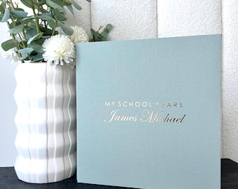 Könnte beinhalten: Ein hellblaues, quadratisches Buch mit dem Titel "MY SCHOOL YEARS" und dem Namen "James Michael" in Goldbuchstaben. Das Buch steht neben einer weißen Vase mit weißen Blumen und grünem Laub.