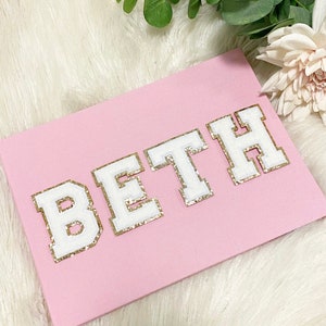 May include: A pink notebook with the name "BETH" spelled out in white letters with gold glitter trim.
