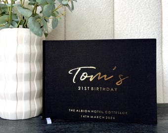 Könnte beinhalten: Ein schwarzes Buch mit goldfarbenen Buchstaben. Der Text lautet "Tom's 21st Birthday", gefolgt von "THE ALBION HOTEL COTTESLOE 14TH MARCH 2026". Links vom Buch steht eine weiße Vase mit Blumen auf dunkler Oberfläche.