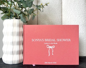Könnte beinhalten: Ein korallenfarbenes Gästebuch für die Brautparty mit dem Text "SONYA'S BRIDAL SHOWER Letters to the Bride" in Weiß. Unter dem Text befindet sich eine weiße Schleifengrafik mit dem Datum "28th March 2026" am unteren Rand. Links steht eine weiße Vase mit weißen Blumen und Grün.