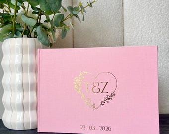 Könnte beinhalten: Ein rosa Gästebuch mit goldfarbenen Buchstaben. Das Cover zeigt ein herzförmiges Design mit den Initialen "T&Z" und dem Datum "22.03.2026". Eine weiße Vase mit weißen Blumen und Grünzeug befindet sich im Hintergrund.