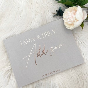 May include: A grey linen guest book with the text "Lara & Billy Addison" in white and gold foil. The date "26 March 2023" is printed in gold foil below the names.