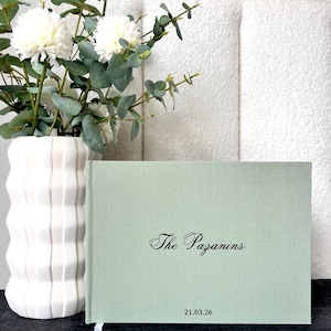 Puede incluir: Un libro de lino verde claro con el texto "The Pazanins" y la fecha "21.03.26" en escritura negra. El libro está sobre una superficie oscura, con un jarrón blanco de flores y un fondo blanco texturizado.