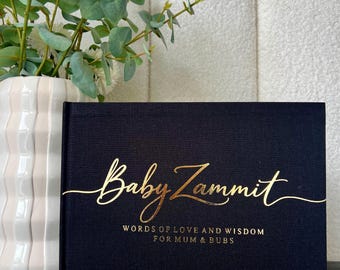 Könnte beinhalten: Ein schwarzes Baby-Erinnerungsbuch mit goldfarbenem Schriftzug "Baby Zammit" und "Words of Love and Wisdom for Mum & Bubs". Das Buch steht neben einer weißen Vase mit weißen Blumen und Grün.
