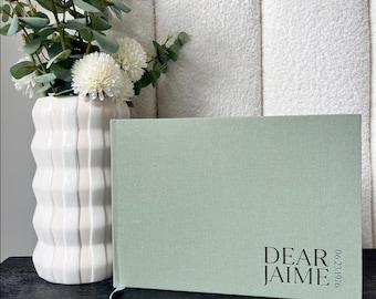 May include: A sage green hardcover journal with the text "DEAR JAIME" printed in black. A white ribbon bookmark is visible. The journal is next to a white vase with white flowers and greenery.