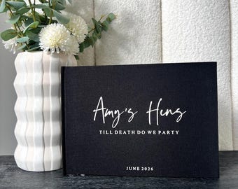 Könnte beinhalten: Ein schwarzes Buch mit weißer Schrift, das "Amy's Hens" und "Till Death Do We Party" sowie "June 2026" darunter zeigt. Links vom Buch befindet sich eine weiße Vase mit weißen Blumen und Grünzeug. Der Hintergrund ist eine strukturierte weiße Wand.