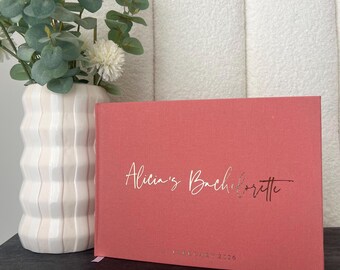 Könnte beinhalten: Ein korallenfarbenes Buch mit dem Text "Alicia's Bachelorette" in goldener Schrift. Das Buch steht neben einer weißen Vase mit weißen und grünen Blumen. Das Datum "1. Januar 2026" ist unten auf dem Buch aufgedruckt.