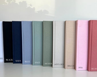 Op de afbeelding: Tien hardcover dagboeken in verschillende kleuren, waaronder wit, zwart, marineblauw, blauw, salie, jagergroen, lichtgrijs, biscuit, roze en blush roze. Elk dagboek heeft een linnen omslag en de kleurnaam is in wit gedrukt aan de onderkant van de rug.