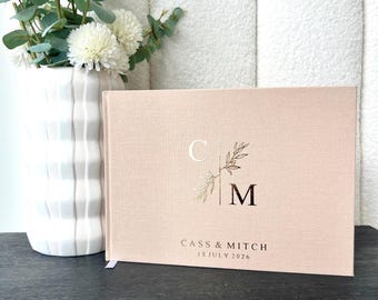 Könnte beinhalten: Ein hellrosa Hochzeitsgästebuch mit goldfarbenen Buchstaben und floralen Akzenten. Auf dem Einband stehen die Initialen "C" und "M", die Namen "Cass & Mitch" und das Datum "18. Juli 2026". Eine weiße Vase mit Blumen ist im Hintergrund.