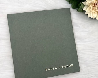 May include: A dark green square notebook with the text "BALI & LOMBOK" printed in gold on the front cover.