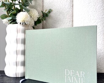 Könnte beinhalten: Hellgrünes Hardcover-Journal mit dem weißen Text "DEAR JAIME". Das Journal liegt auf einer dunklen Oberfläche neben einer weißen Vase mit weißen Blumen und Grünzeug. Im Hintergrund befindet sich ein weißes, strukturiertes Kopfteil.