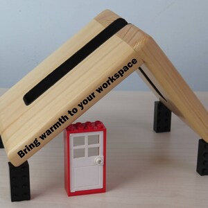 K&ouml;nnte beinhalten: Ein Laptopst&auml;nder aus Holz mit schwarzen Gummif&uuml;&szlig;en und einem schwarzen Gummistreifen an der Oberseite. Der St&auml;nder ist nach oben geneigt und hat den Text "Bring warmth to your workspace" auf der Seite aufgedruckt. Ein roter Lego-Stein mit einer wei&szlig;en T&uuml;r befindet sich im Vordergrund.
