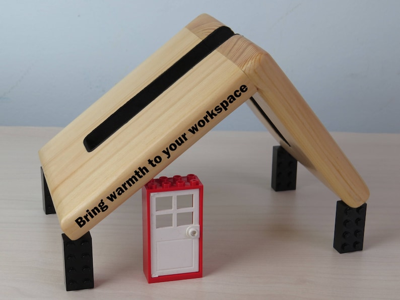 K&ouml;nnte beinhalten: Ein Laptopst&auml;nder aus Holz mit schwarzen Gummif&uuml;&szlig;en und einem schwarzen Gummistreifen an der Oberseite. Der St&auml;nder ist nach oben geneigt und hat den Text "Bring warmth to your workspace" auf der Seite aufgedruckt. Ein roter Lego-Stein mit einer wei&szlig;en T&uuml;r befindet sich im Vordergrund.