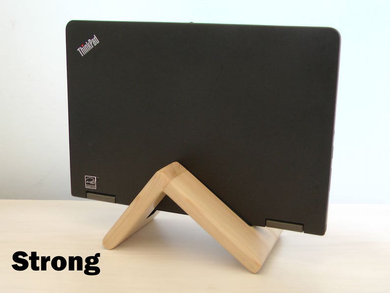 K&ouml;nnte beinhalten: Ein schwarzer Laptop mit dem ThinkPad-Logo auf dem Deckel liegt auf einem Holzst&auml;nder. Der St&auml;nder besteht aus zwei V-f&ouml;rmigen Holzst&uuml;cken. Der Laptop steht auf einer hellen Holzoberfl&auml;che. Das Wort "Strong" ist in schwarzen Buchstaben auf der Oberfl&auml;che geschrieben.