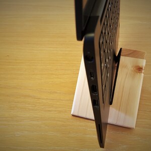 K&ouml;nnte beinhalten: Ein schwarzer Laptop steht auf einem Holzst&auml;nder. Der Laptop ist geschlossen und der St&auml;nder ist aus Naturholz.
