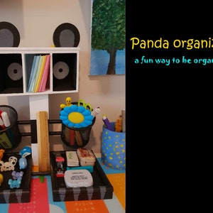 Könnte beinhalten: Ein pandaförmiger Schreibtisch-Organizer mit schwarzen und weißen Ohren, einem weißen Körper und schwarzen Augen. Der Organizer hat drei Fächer zum Verstauen von Gegenständen, zwei Netzstifthalter und ein Lineal aus Holz. Der Organizer steht auf einem Schreibtisch mit einem schwarz-weißen Zebra-Muster. Der Text "Panda Organizer" und "a fun way to be organized" ist in gelber und blauer Schrift auf schwarzem Hintergrund geschrieben.