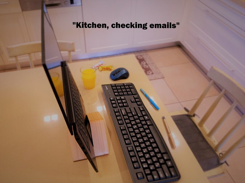 K&ouml;nnte beinhalten: Ein schwarzer Laptop ist auf einem Holztisch mit einer Tastatur, einer Maus und einem Glas Orangensaft ge&ouml;ffnet. Der Laptop ist auf einem Holzst&auml;nder aufgestellt. Der Text "K&uuml;che, E-Mails checken" ist &uuml;ber dem Laptop sichtbar.