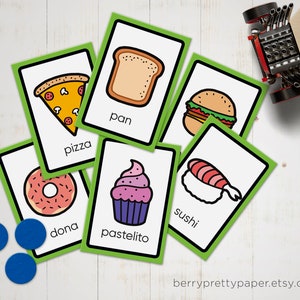 May include: Green flash cards with illustrations of different foods and their names in English and Spanish. The foods include pizza, donut, bread, cupcake, sushi, and a hamburger.